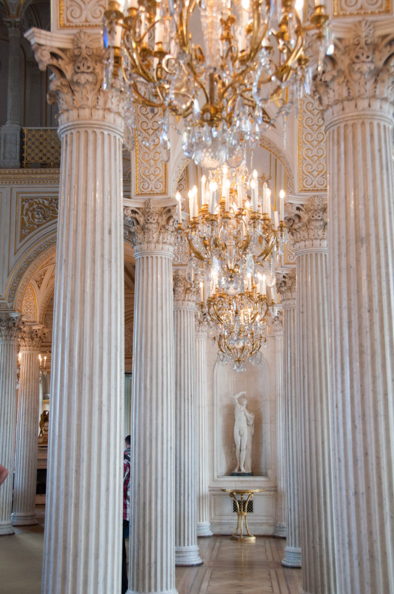White-and-gold Hermitage gallery with fluted columns, crystal chandeliers, ornate ceiling, and classical statue in a far niche.