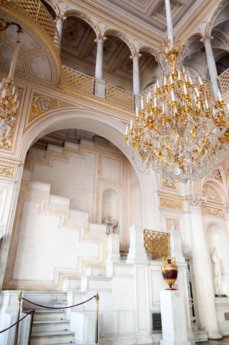 Monumental stepped white architectural feature beneath a crystal chandelier, with upper gallery, sculpture, columns, and gilded urn.