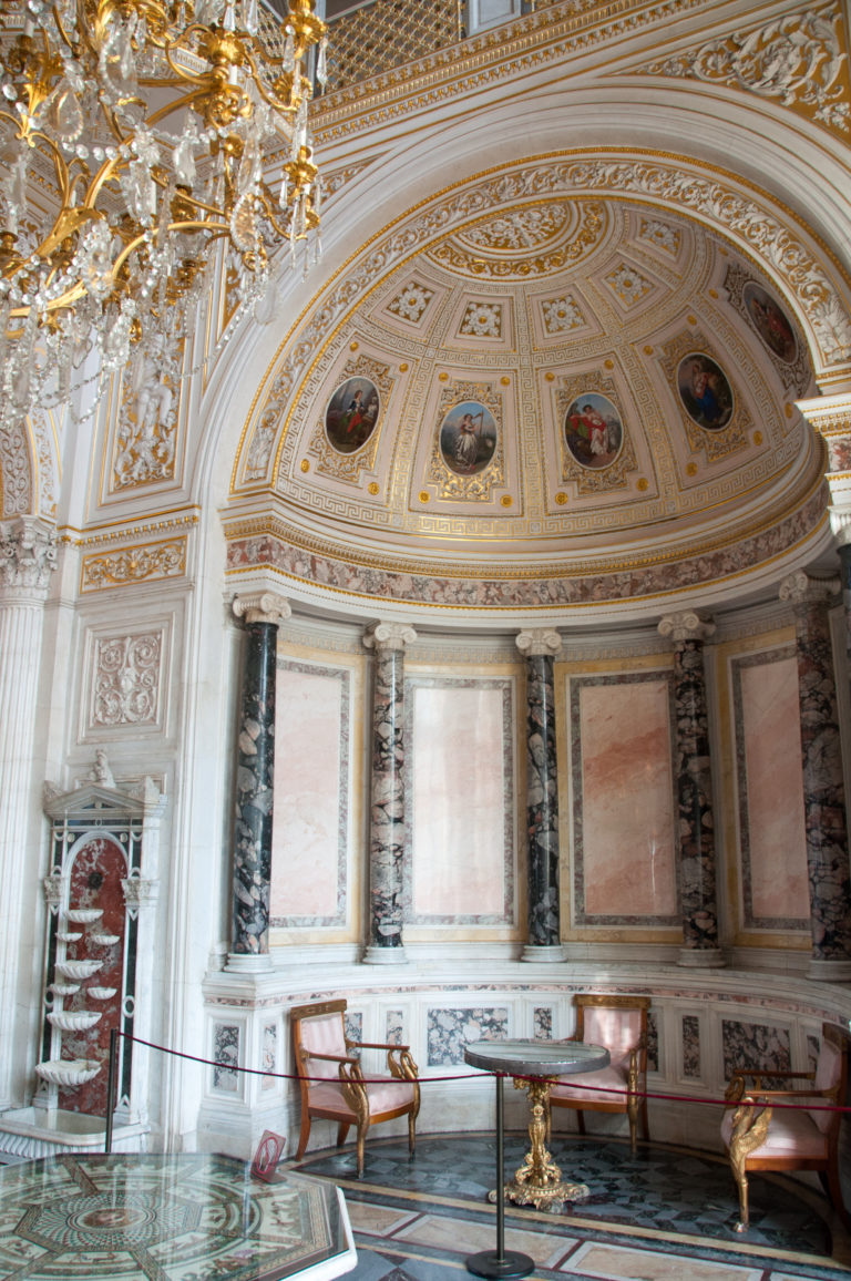 Semicircular Hermitage alcove with multicolored marble panels, dark columns, painted medallions, gilded ornament, and upholstered chairs.