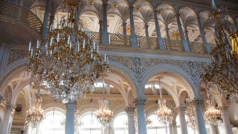 White-and-gold Hermitage hall with repeated arches, slender columns, upper gallery, and multiple crystal chandeliers viewed upward.