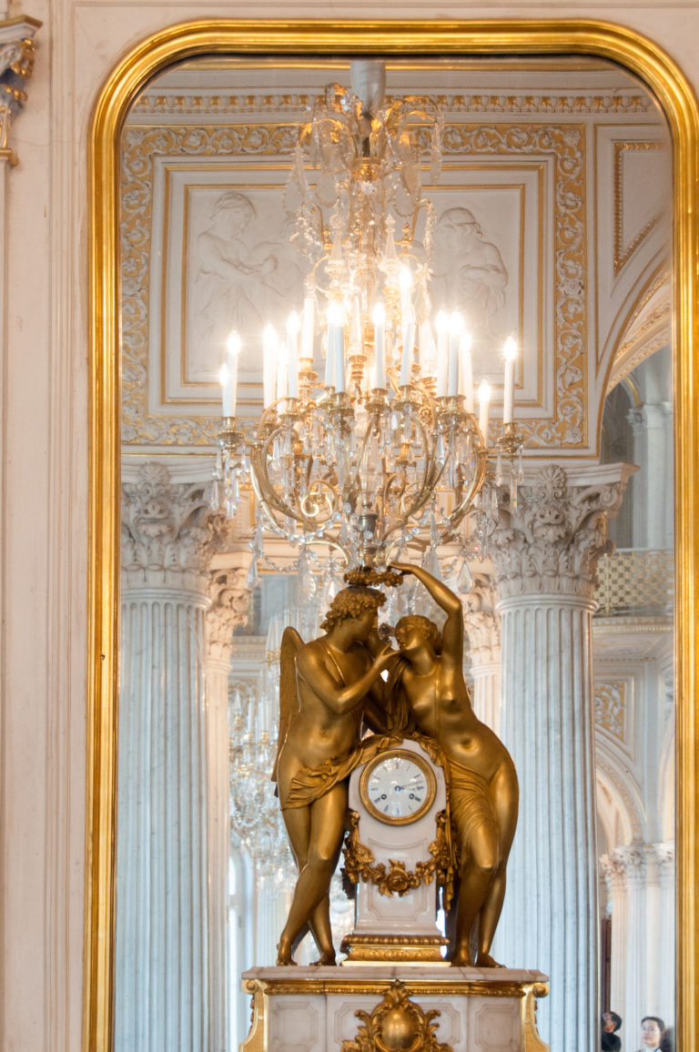 Gilded clock supported by two sculpted elephants with raised trunks, reflected with chandelier and pale columns in a large mirror.