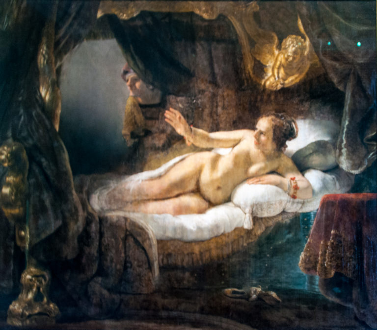 Reclining nude woman on a bed reaching one arm outward, surrounded by heavy curtains and warm golden light.
