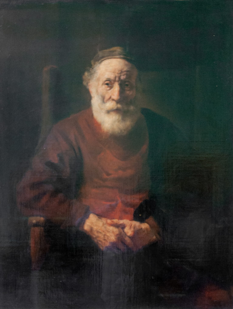 Seated elderly bearded man in a red garment and dark cap, hands folded before him against a shadowed background.