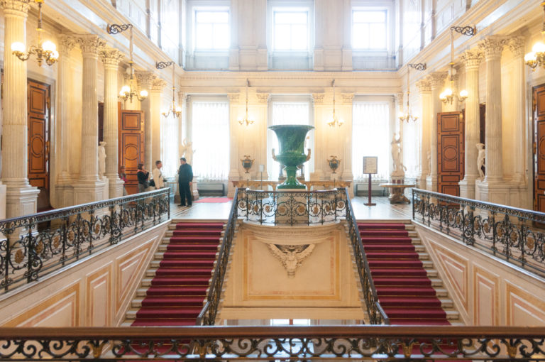 Symmetrical red-carpeted Hermitage staircase with dark balustrades, monumental green vase at the upper landing, and visitors near tall windows.
