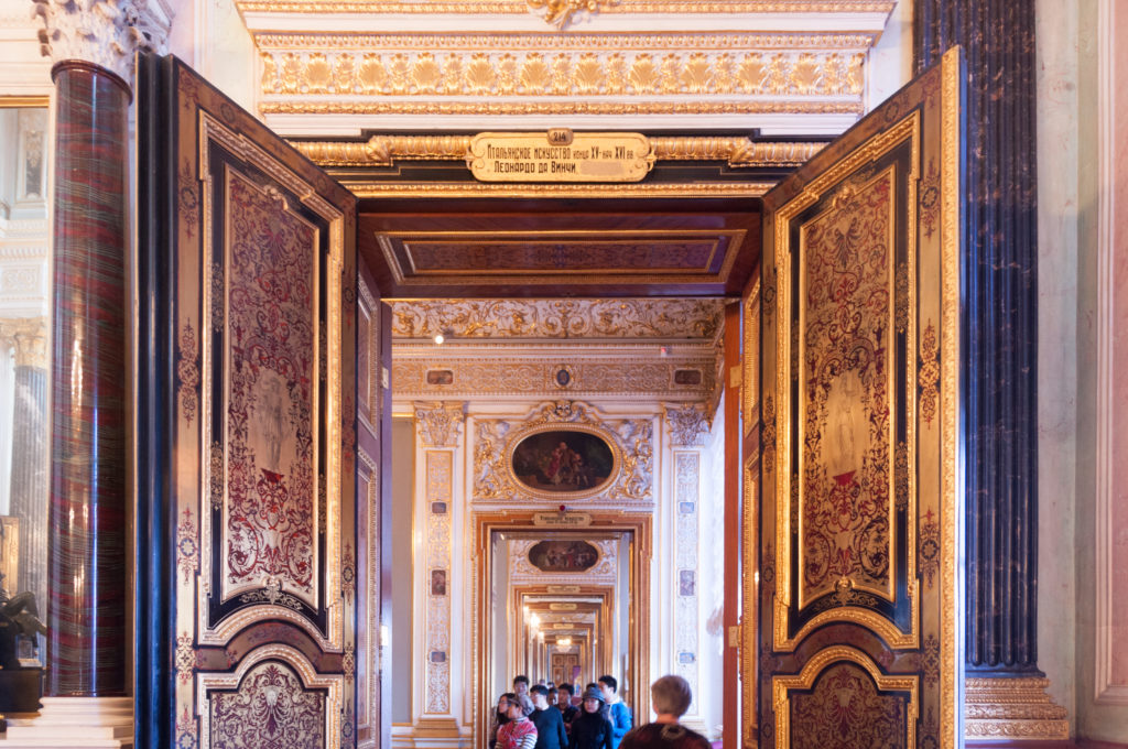 Long Hermitage enfilade through open ornate doors, with dark columns, gilded moldings, painted panels, and visitors in the distance.