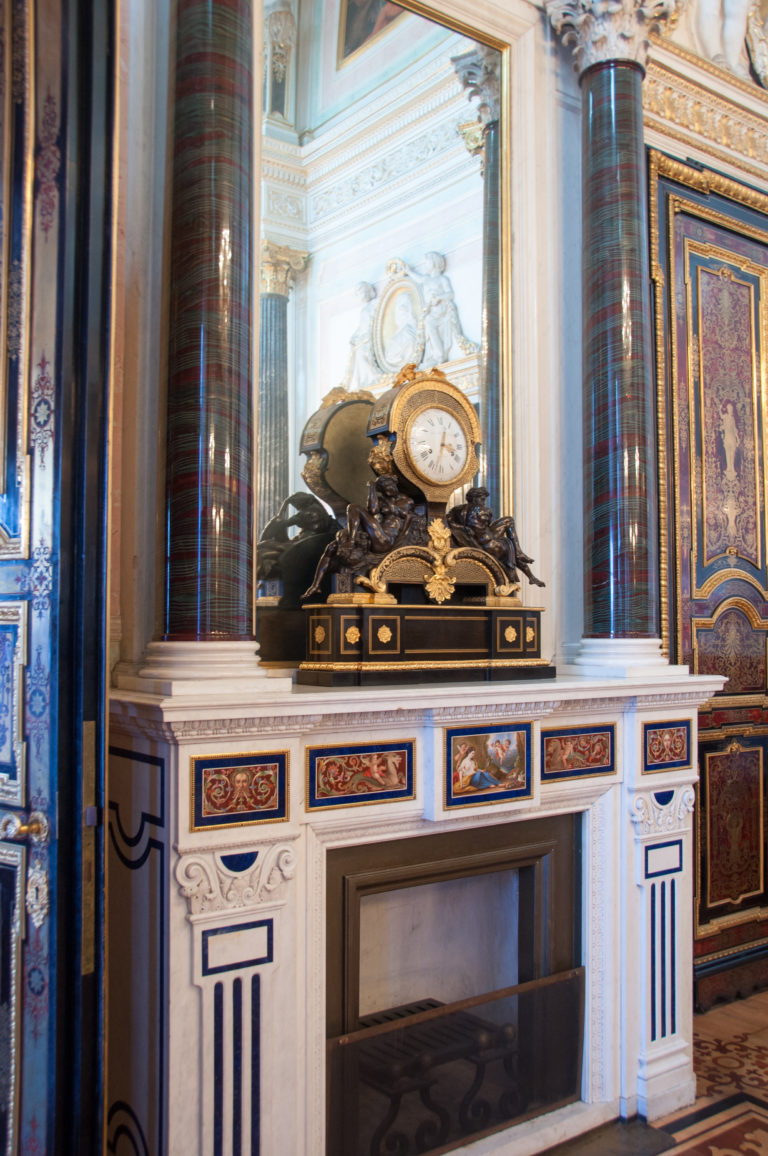 Dark ornamental mantel clock with circular dial and sculptural base above a pale fireplace between polished columns.
