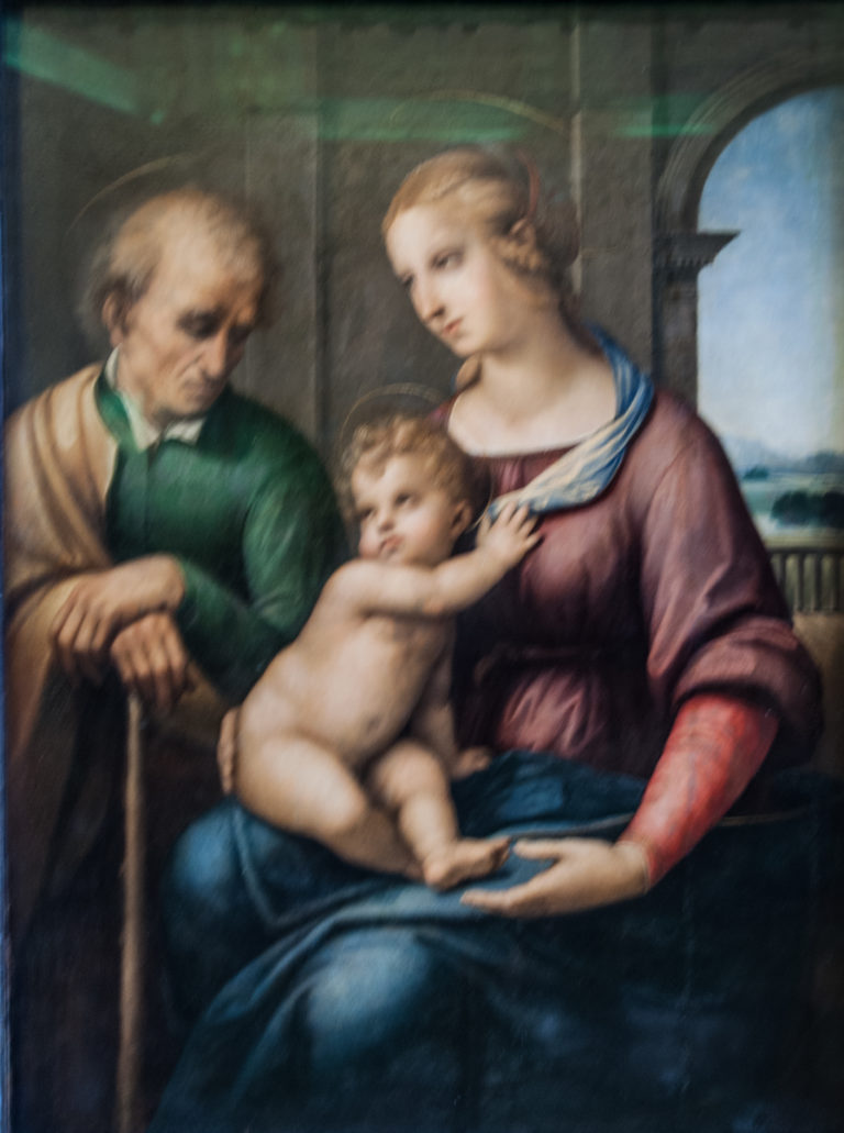 Virgin with Christ Child and elderly bearded man grouped closely before a dark interior and distant landscape.