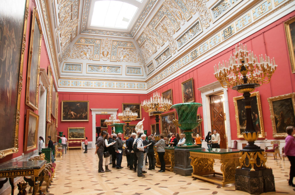 Red-walled Hermitage gallery with large paintings, crystal chandeliers, ornate ceiling, green monumental vessels, and groups of visitors.