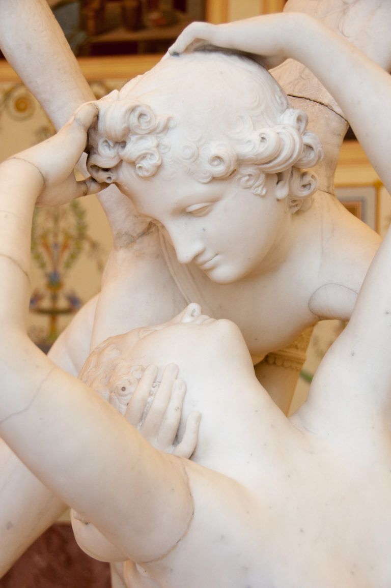 Close-up of two pale marble figures embracing with interlocked arms, carved curls, and faces turned closely toward one another.