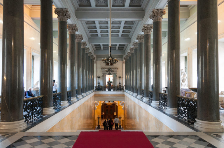 Symmetrical Hermitage upper landing with dark stone columns, coffered ceiling, ornate balustrades, red carpet, and stair opening.