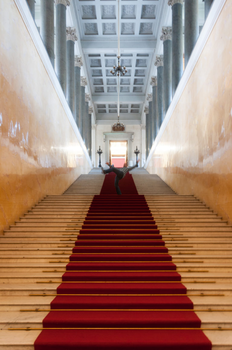 Broad pale staircase rising beneath dark columns and a coffered ceiling, with a narrow red carpet runner centered on the steps.