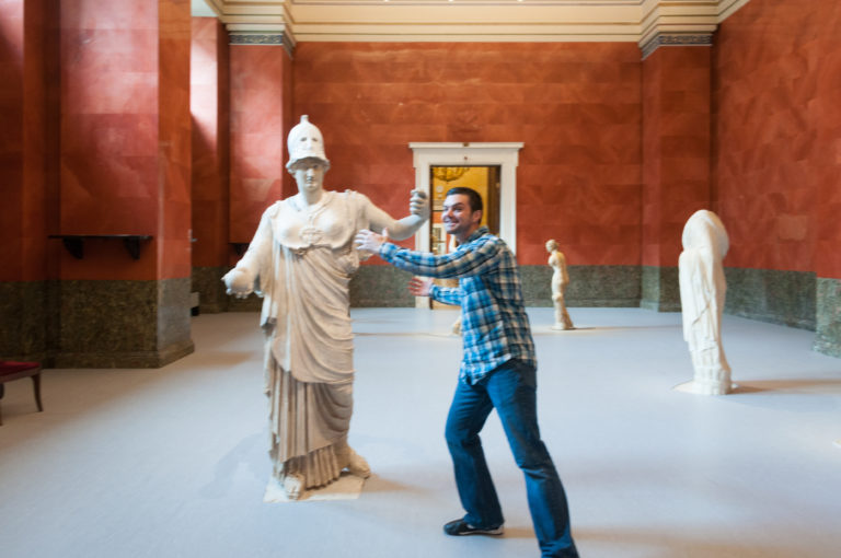 Visitor in plaid shirt posing beside a pale helmeted classical statue with outstretched arm in a red-walled gallery.
