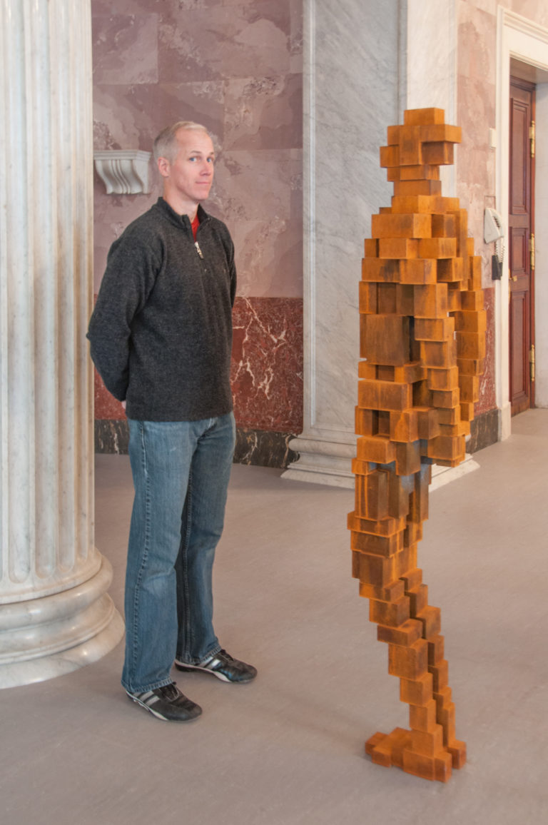 Visitor standing beside a human-like sculpture built from stacked rectangular wooden blocks in a marble museum interior.