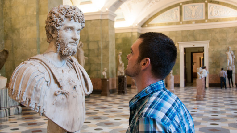 Visitor in plaid shirt facing a bearded marble portrait bust in a large sculpture gallery with patterned floor.