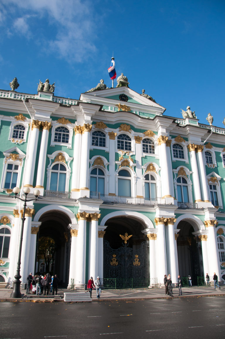Green-and-white Winter Palace façade with white columns, gilded details, arched windows, central gateway, and visitors on Palace Square.
