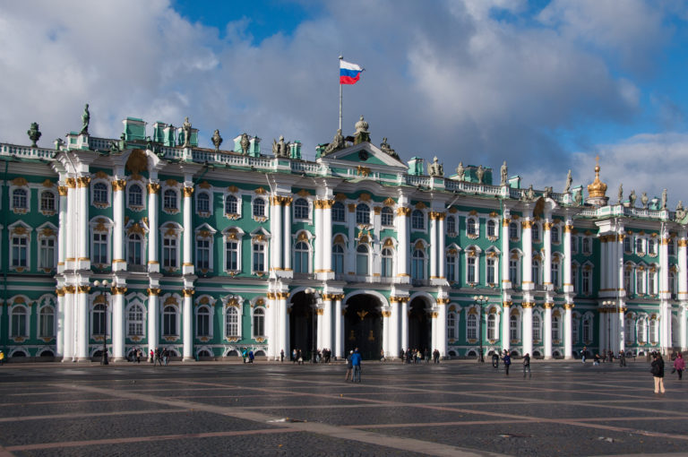 Wide Winter Palace façade across Palace Square, with green walls, white columns, rooftop statues, Russian flag, and scattered pedestrians.