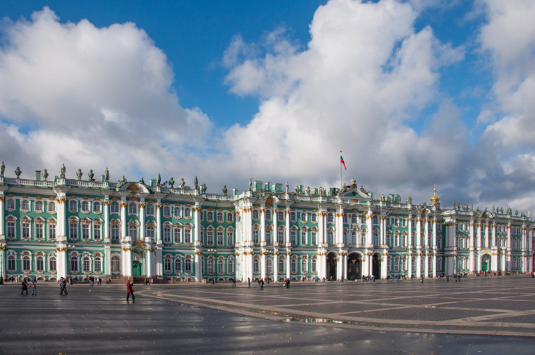 Panoramic Winter Palace façade under large white clouds, with green walls, white columns, rooftop statues, and broad Palace Square foreground.