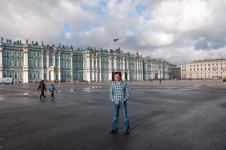 Visitor in plaid shirt standing on Palace Square with the green-and-white Winter Palace and Russian flag in the background.