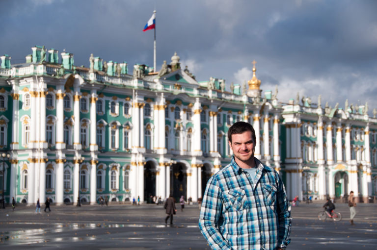 Waist-up portrait of a visitor on Palace Square with the Winter Palace, gilded details, rooftop statues, and Russian flag behind.