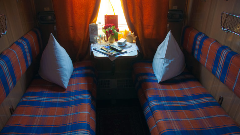 Two facing plaid train berths with white pillows and a small table beneath a curtained window.