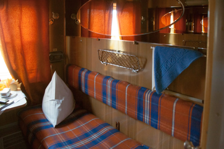 Plaid upholstered train berth with white pillow in a narrow wood-paneled sleeping compartment with mirror and curtain.