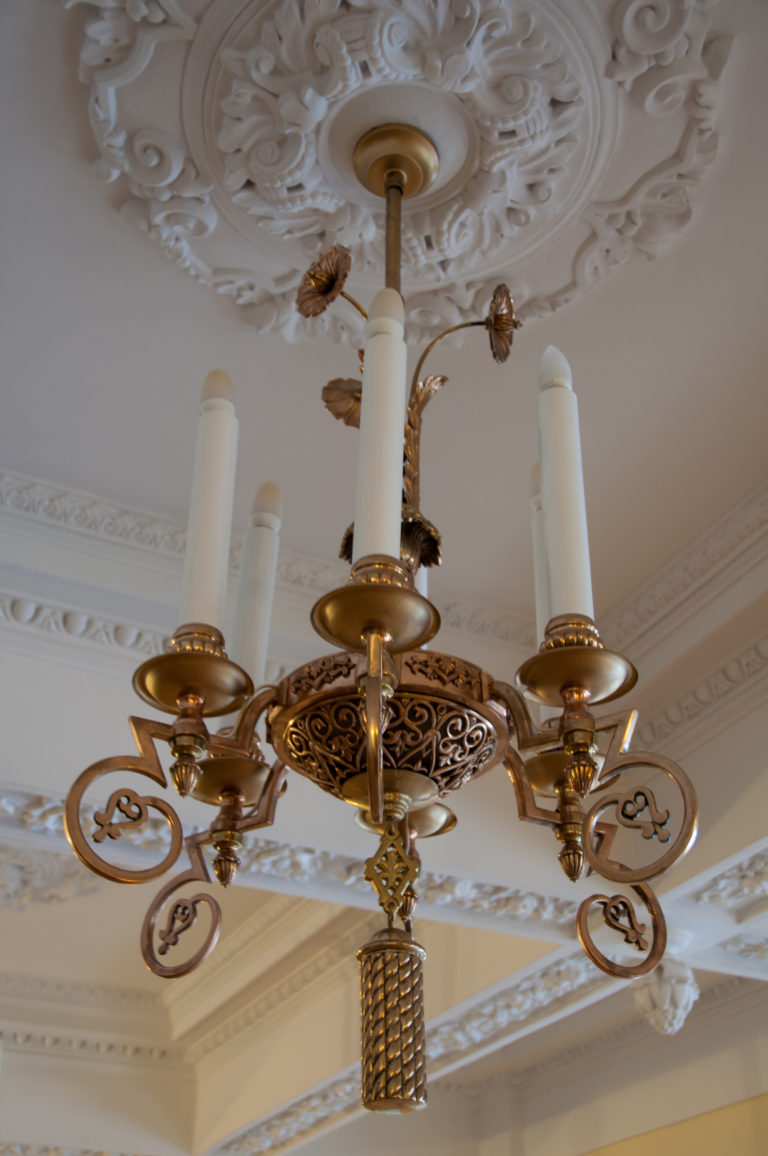 Small ornate chandelier with candle-form lamps beneath a white molded plaster ceiling and ceiling rosette.