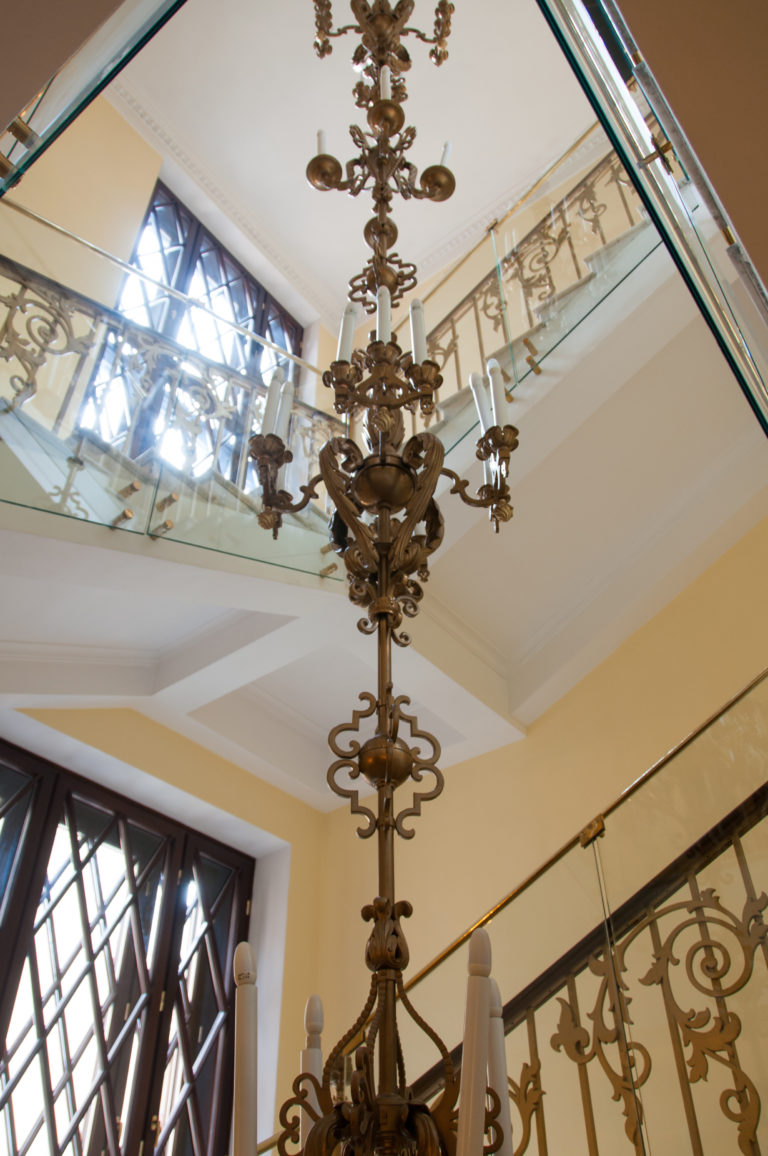 Tall ornate metal fixture hanging through a multi-level stairwell beneath a glazed roof and decorative railings.
