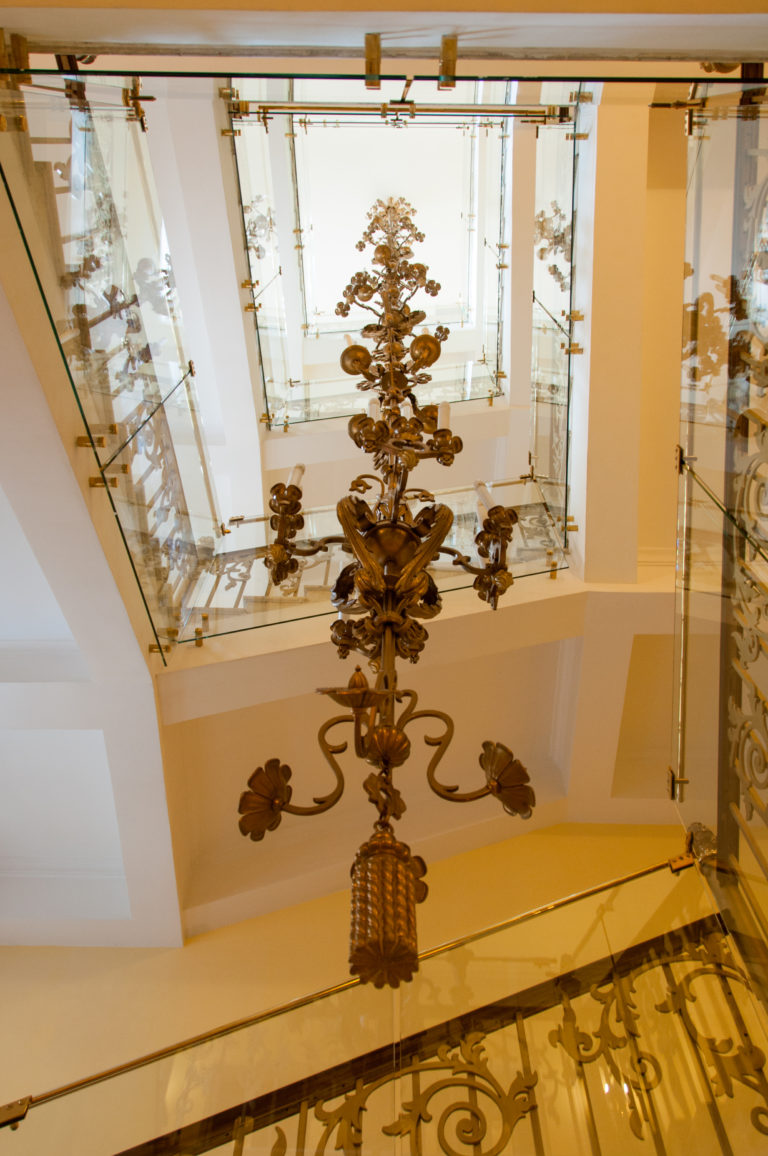 Long ornate metal hanging fixture descending beneath a glazed atrium ceiling with decorative railings around it.