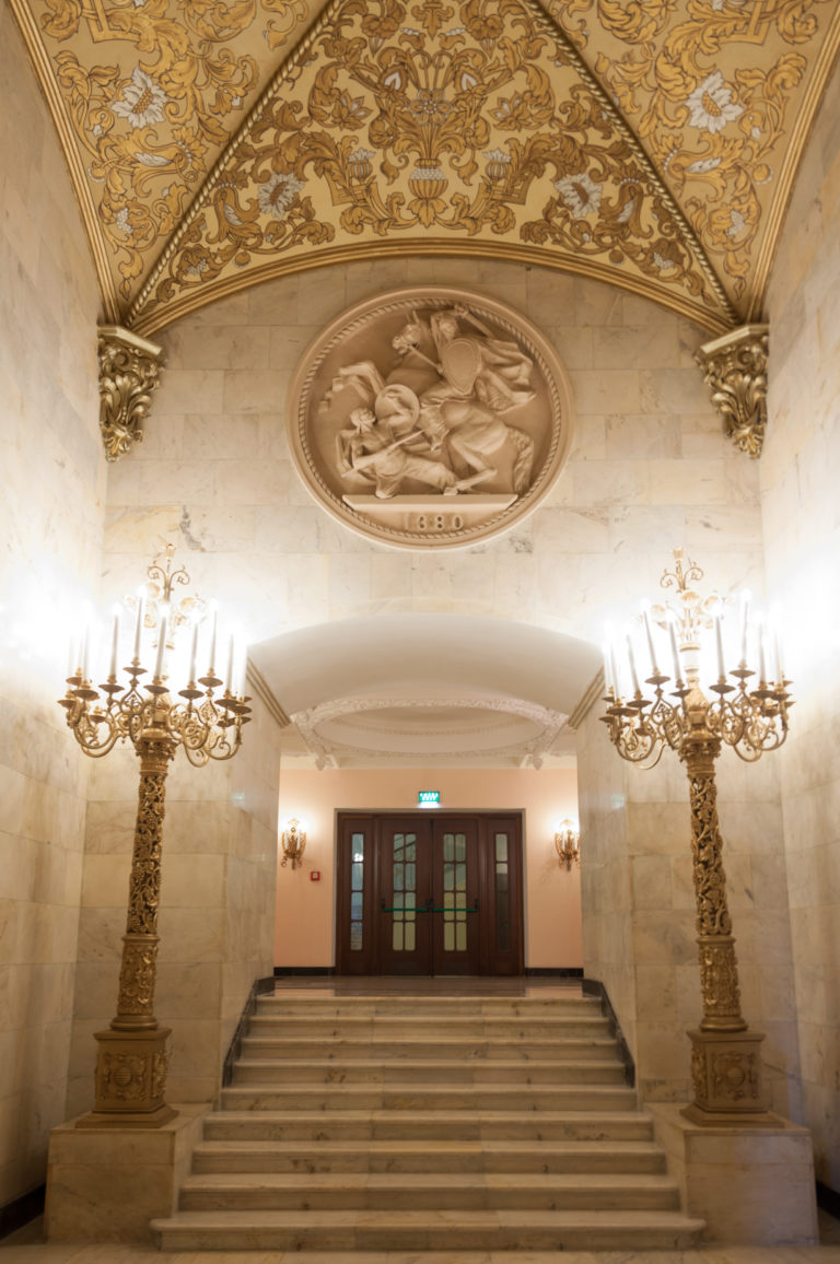 Marble stairs leading to double doors between two ornate candelabra, with a circular relief above.