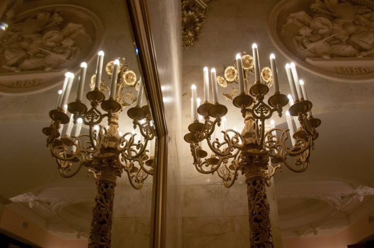Ornate standing candelabrum with candle-form lamps reflected in a tall mirror beneath a molded ceiling.