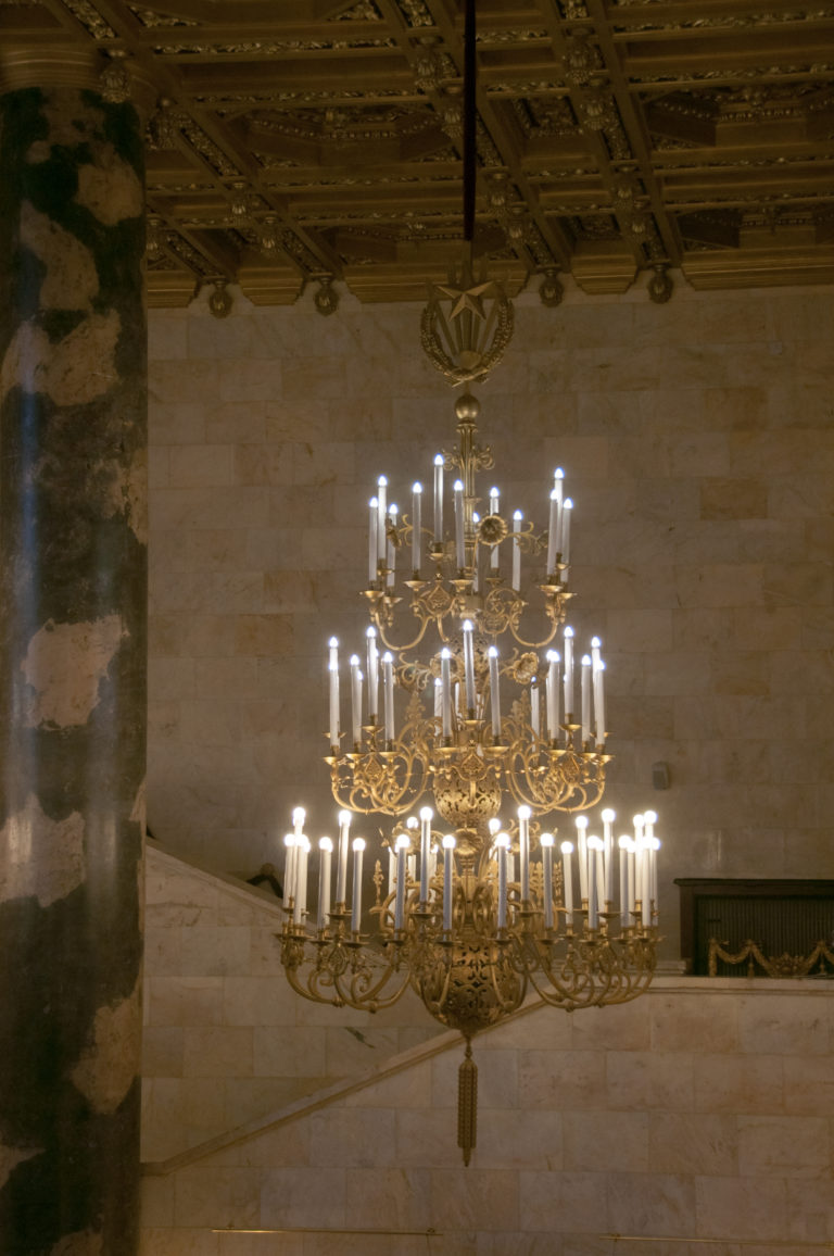 Wide multi-tier chandelier glowing against pale stone walls beside a dark veined column.
