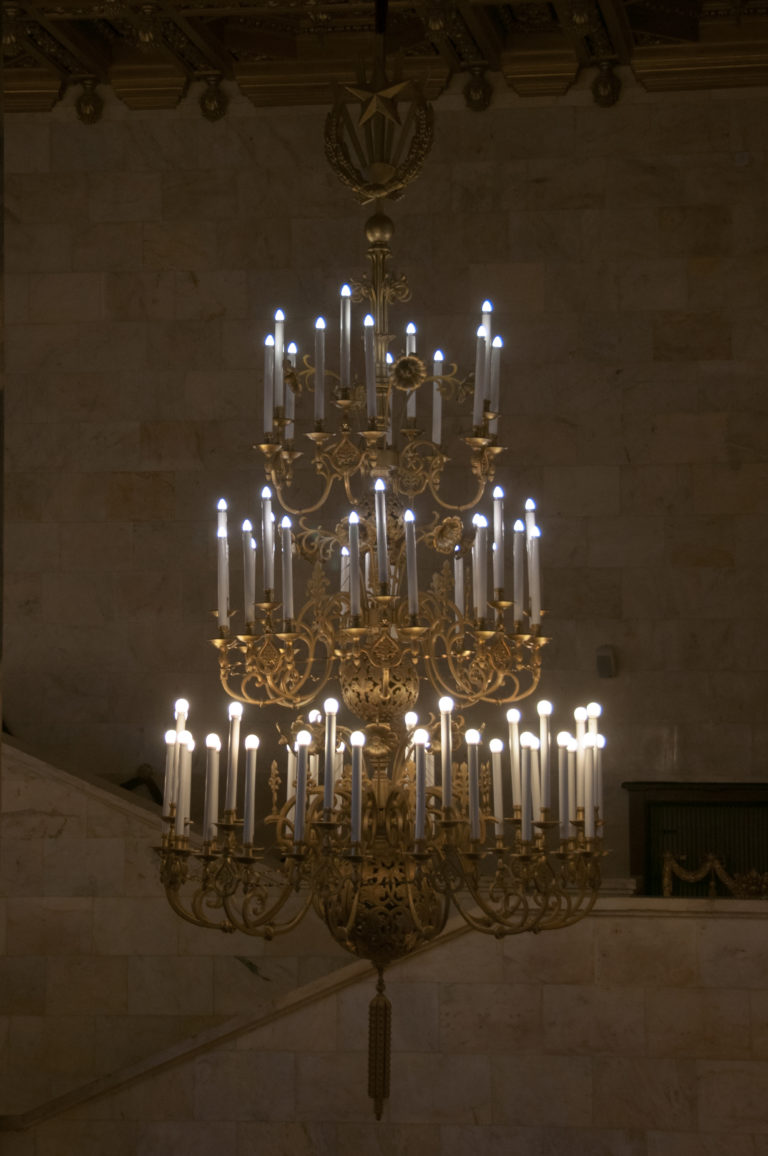 Large illuminated chandelier with several tiers of candle-form lamps hanging against a pale stone wall.