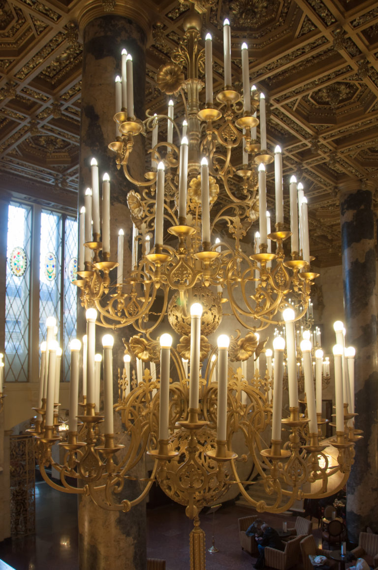 Close view of an ornate gilded chandelier with many candle-form lamps beside a stained-glass window.