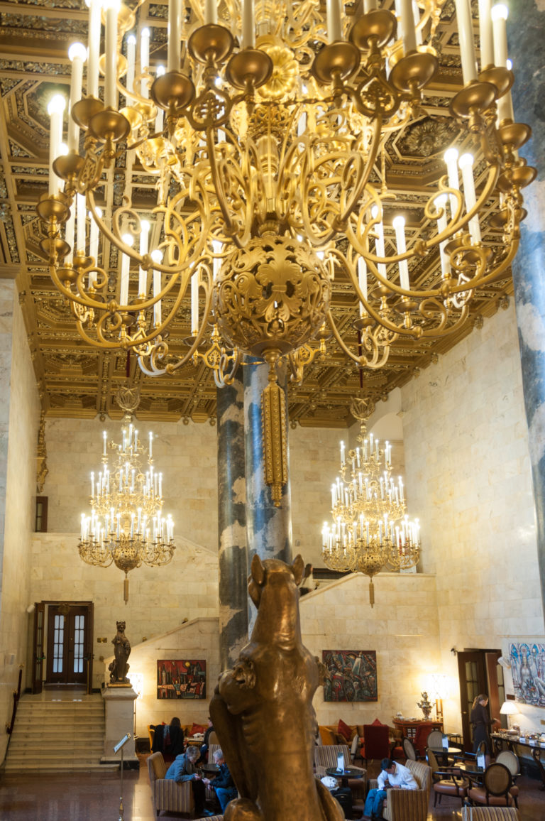 Gilded chandelier above a dark sculptural column in a marble hall with more chandeliers, stairs, seating, and artworks.