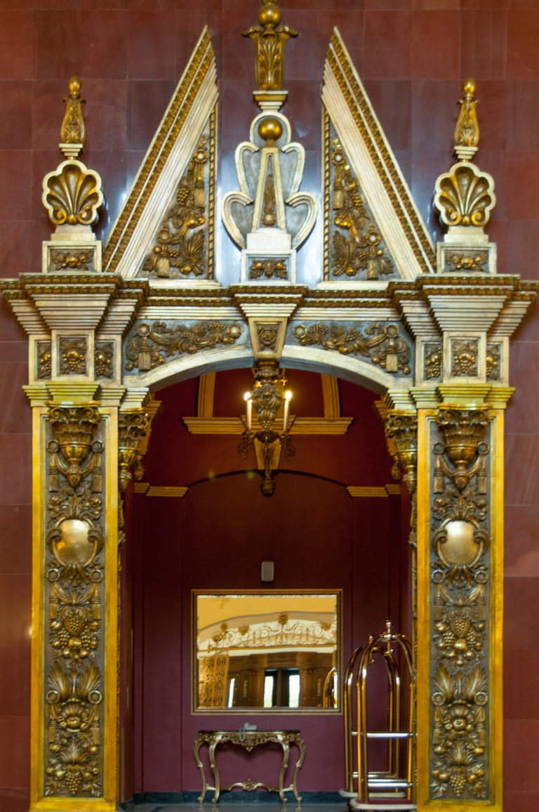 Elaborate gilded doorway with sculptural relief panels and triangular ornament set against deep red walls.