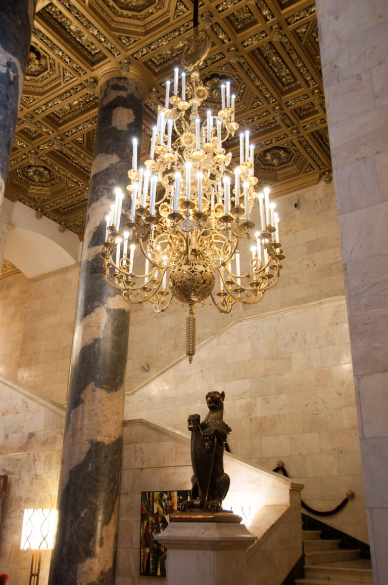 Large gilded chandelier hanging beside a dark marble column beneath an ornate coffered ceiling in a pale stone hall.