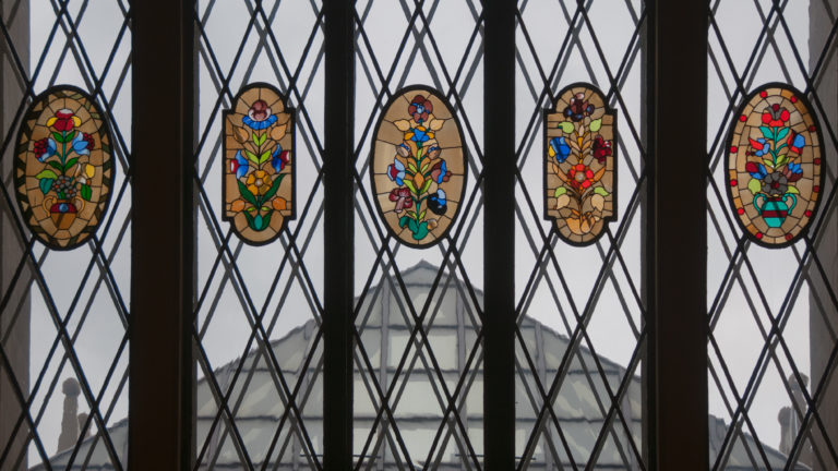 Leaded window with diamond panes and colorful oval floral stained-glass medallions overlooking a hazy city view.
