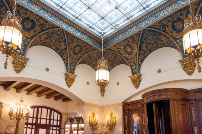 Ornate interior with blue-and-gold vaulted decoration, white arches, hanging lanterns, dark wood doors, and a glazed ceiling.
