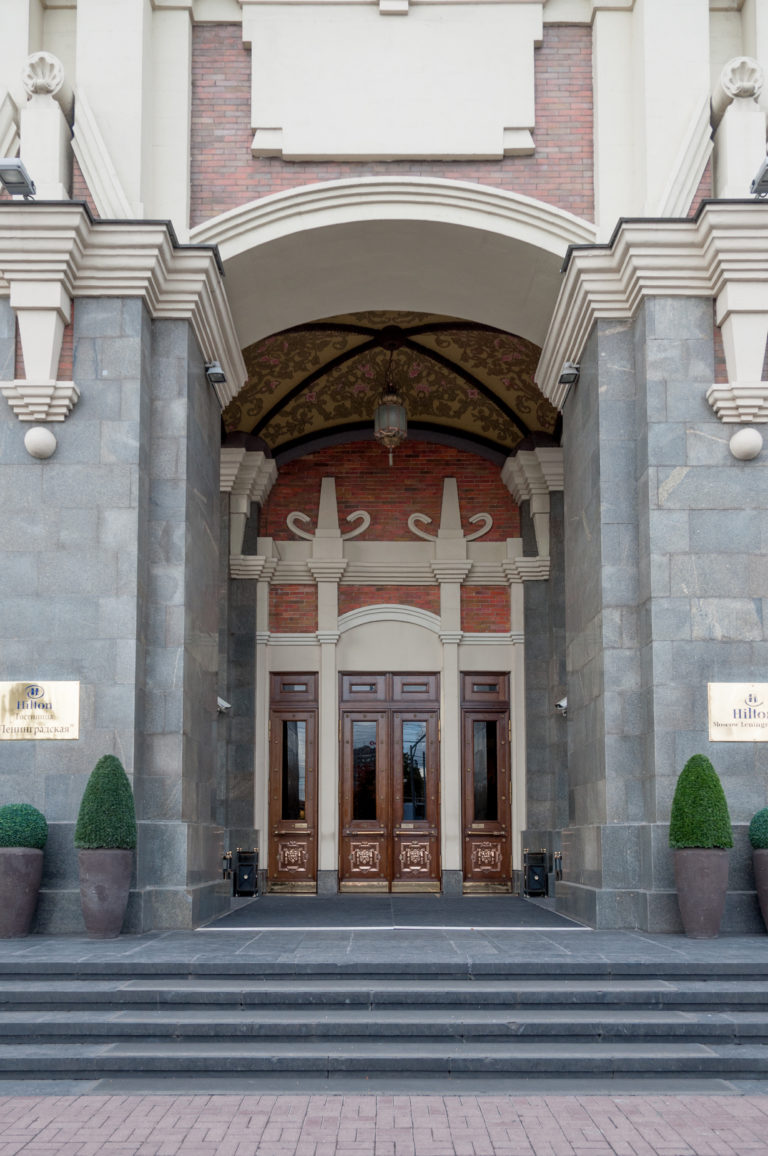Broad steps leading to Hotel National Moscow's recessed entrance with dark stone piers, wooden doors, and paired topiary planters.