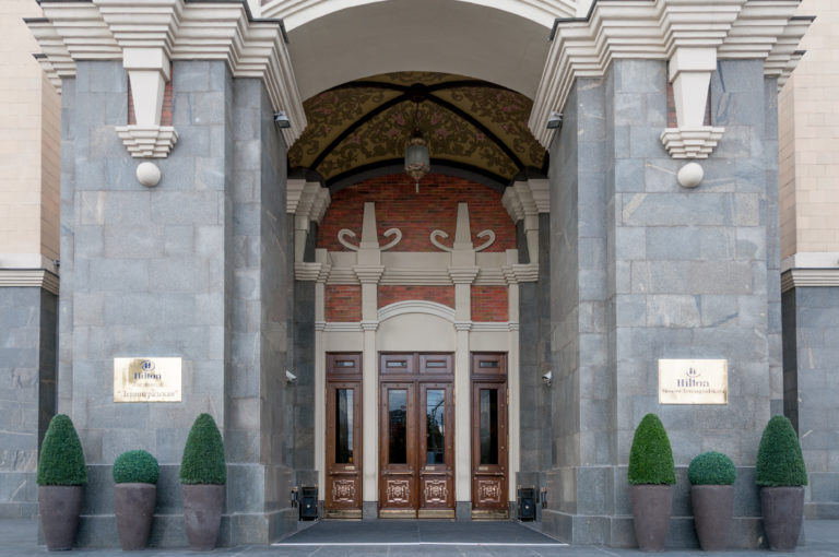 Frontal close view of Hotel National Moscow entrance with wooden doors, dark stone piers, arched decoration, and potted shrubs.