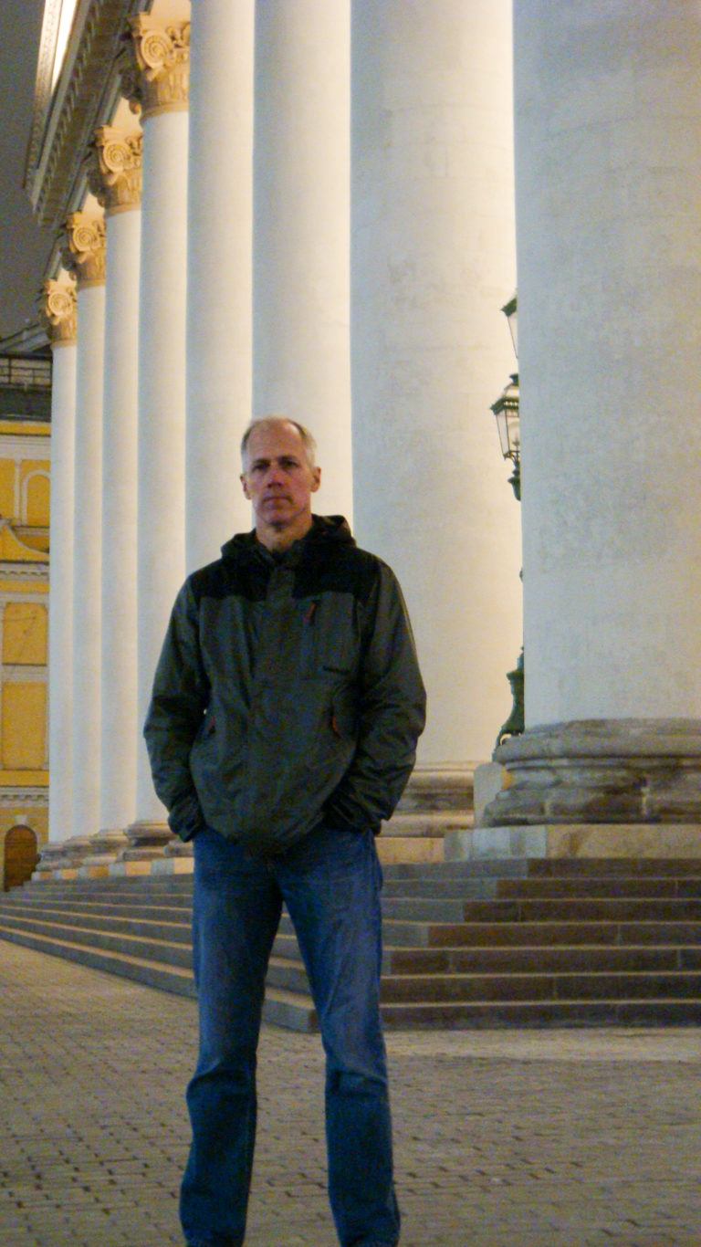 Man standing on broad stone steps between massive white classical columns in Moscow.