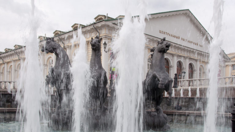 Tall fountain jets and dark horse sculptures in front of a pale building marked with Cyrillic lettering for the Central Exhibition Hall Manege.