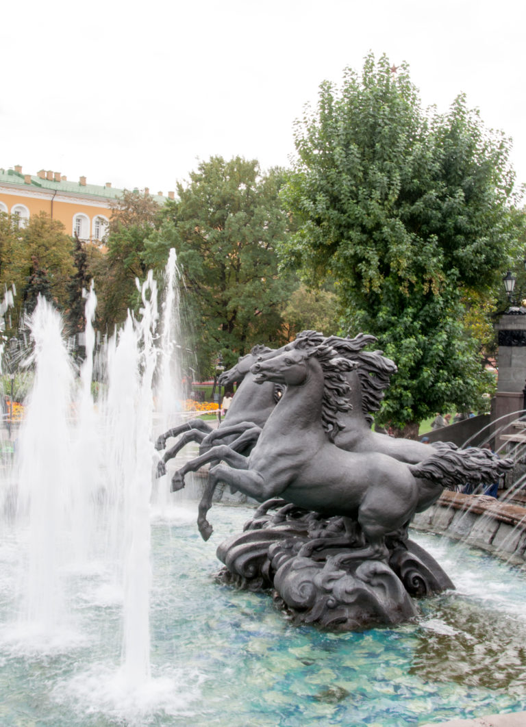 Dark bronze rearing horses surrounded by tall white water jets in a landscaped Moscow fountain.