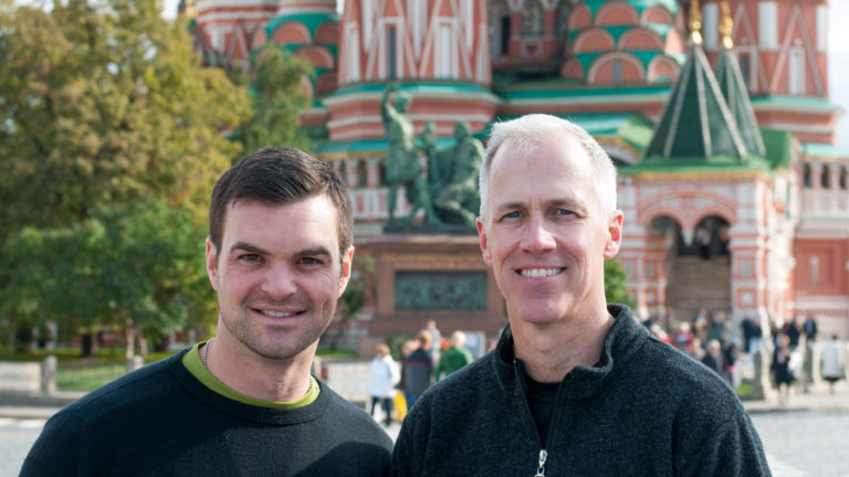 Two men posing outdoors with the colorful towers and onion domes of Saint Basil's Cathedral behind them.