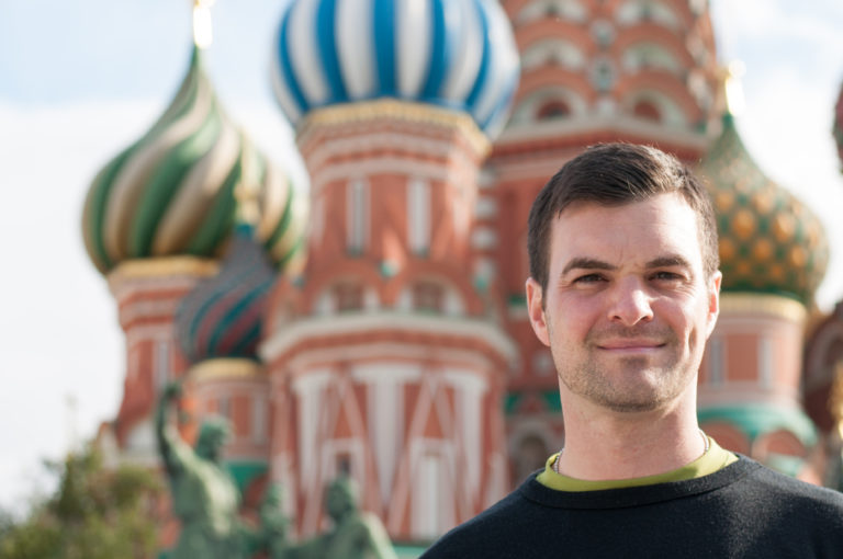Man smiling in the foreground with the colorful onion domes of Saint Basil's Cathedral blurred behind him.