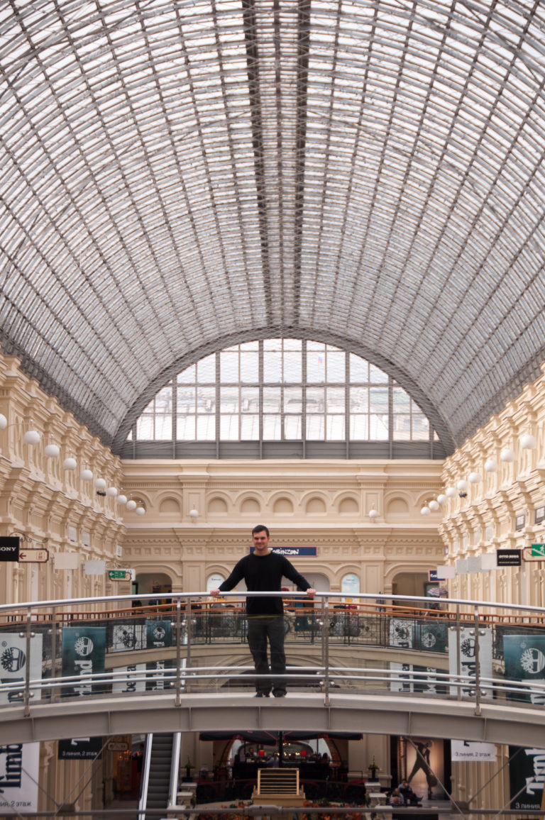 Person standing on an upper bridge inside GUM beneath the expansive gridded glass-and-metal roof.