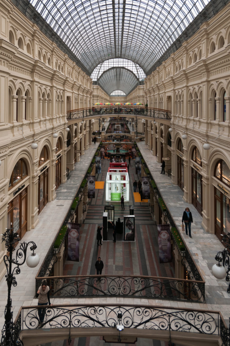 Long interior arcade of GUM beneath a vaulted glass roof, viewed from an upper gallery with shoppers below.