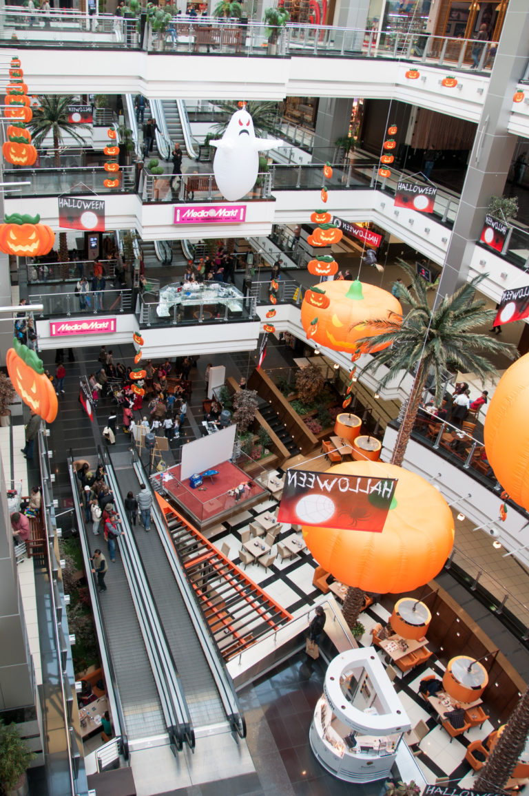 Multi-level shopping mall atrium with escalators, glass railings, storefronts, and large orange hanging decorations.