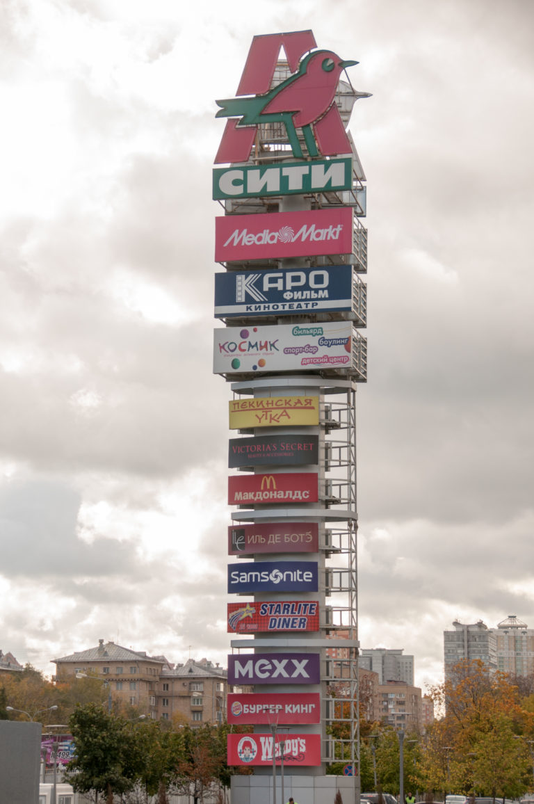 Tall shopping-center directory sign with stacked retail logos against an overcast Moscow sky.