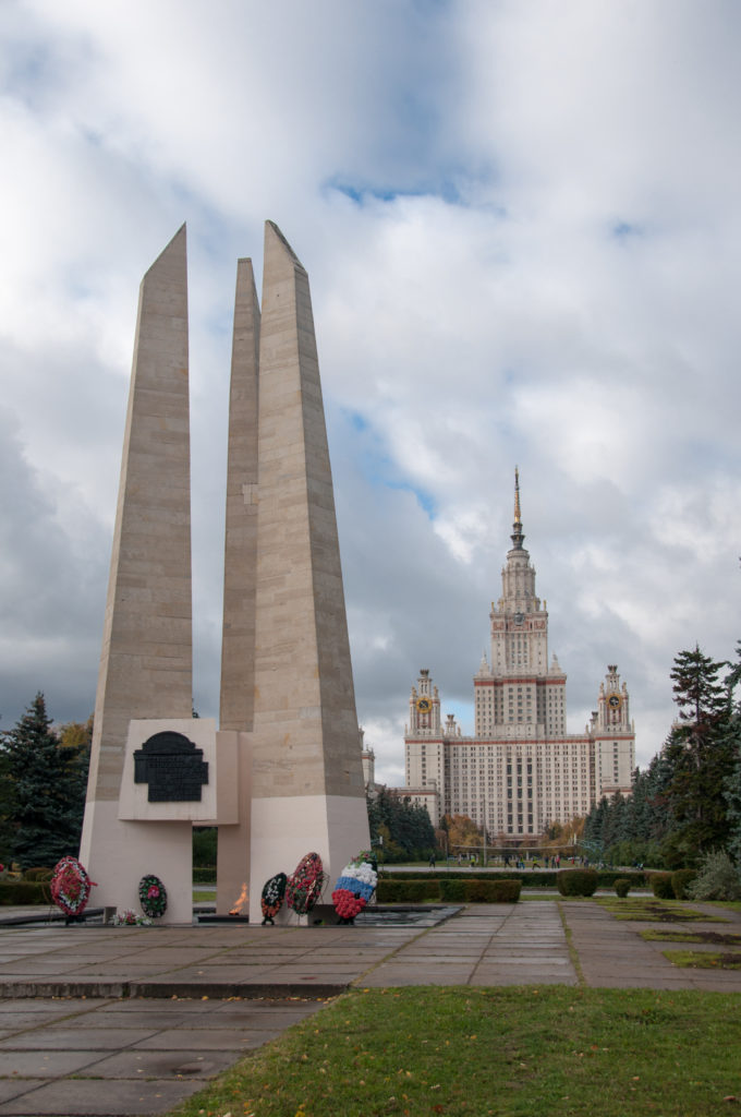 Three tall pale stone memorial slabs with wreaths at their base and Moscow State University centered in the distance.
